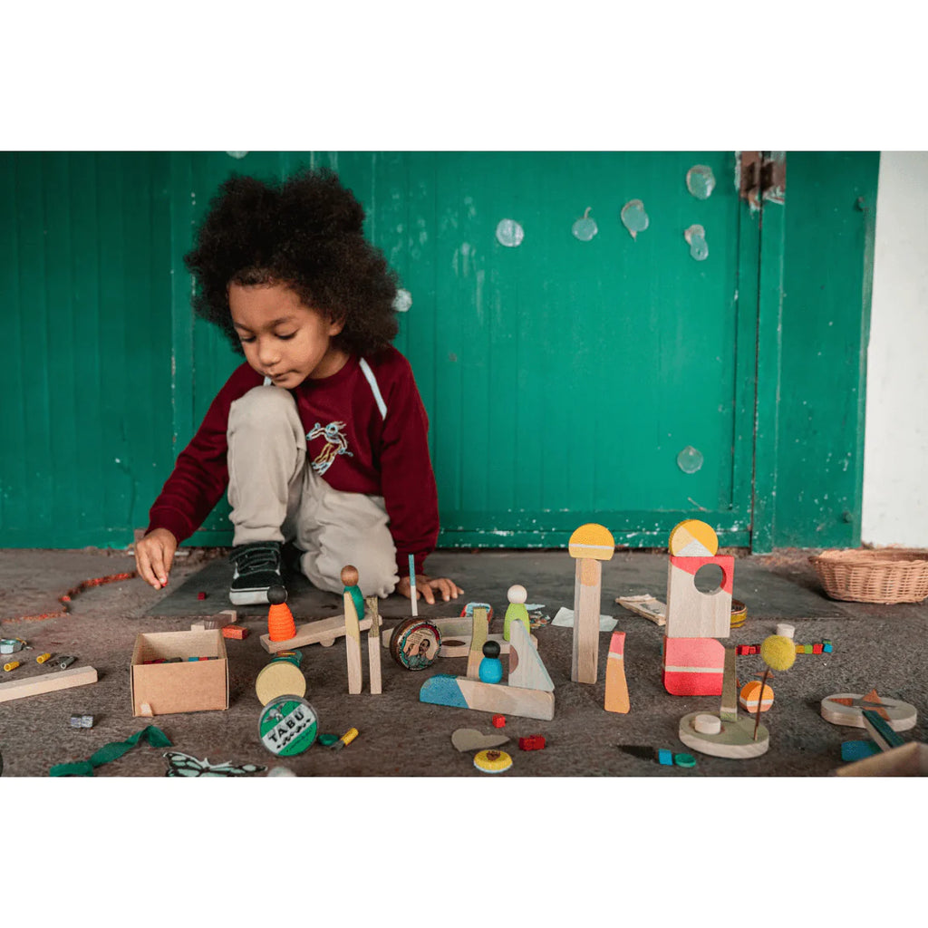 A young child with curly hair is playing with Grapat Happy Place, made from sustainable wood, on a grey floor, in front of a bright green door.
