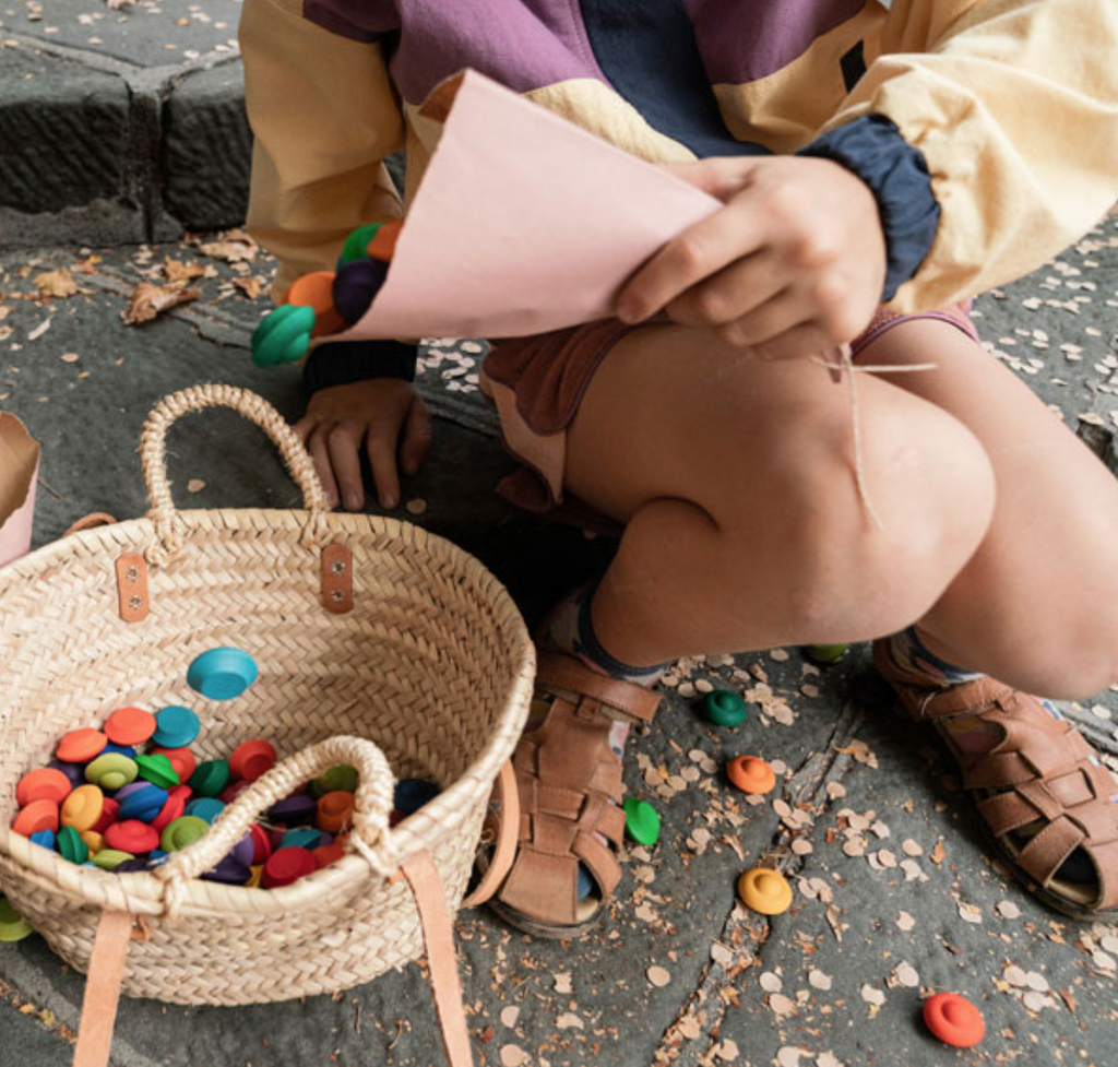 A child in sandals is sitting on the ground, stringing colorful Grapat Mandala Rainbow Flowers. A wicker basket next to them spills assorted beads onto the pavement.