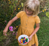A young girl in a yellow dress collects colorful Grapat Mandala Rainbow Mushrooms in a white bowl, standing in a garden surrounded by greenery and flowers.