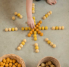 A child's hand arranging small, round orange and yellow Grapat Mandala Honeycomb toys into a sun-like pattern on a concrete floor, with baskets of similar toys nearby.