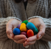 A person wearing a beige knitted sweater holds a collection of brightly colored Grapat Mandala Rainbow Eggs in their cupped hands, clearly displaying the vibrant red, blue, green, yellow, and orange eggs dyed with.
