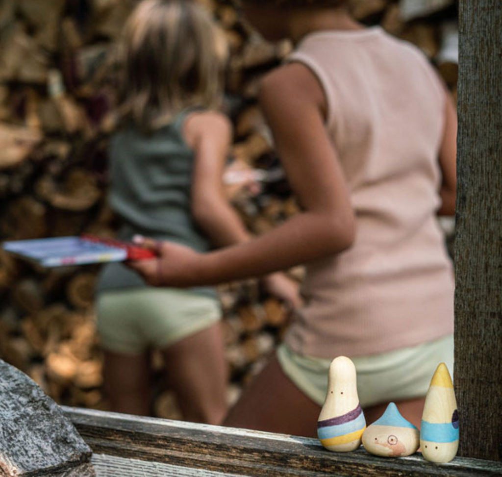 Two children engage in imaginative play near a wood stack, one holds a toy canoe, viewed from behind. In the foreground, two small painted Grapat Wow! Play Sets are perched on a ledge.