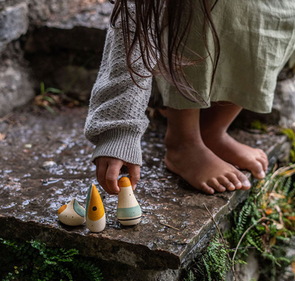 A child in a green skirt and knit sweater sets up tiny, colorful Grapat Hooray! Play Set toys on a mossy stone step, focusing intently on arranging them. Only their lower half and hands are visible.