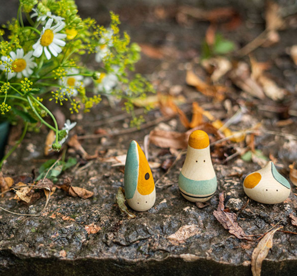 Three Grapat Hooray! Play Set wooden figures with simple, painted features, resembling a small family, placed on a forest floor next to white and yellow flowers.