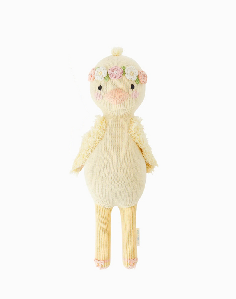 A Cuddle + Kind Duckling plush knitted doll with a pale yellow body and cream-colored wings. It wears a flower crown on its head with green leaves and pink flowers, and a small pink blush on.