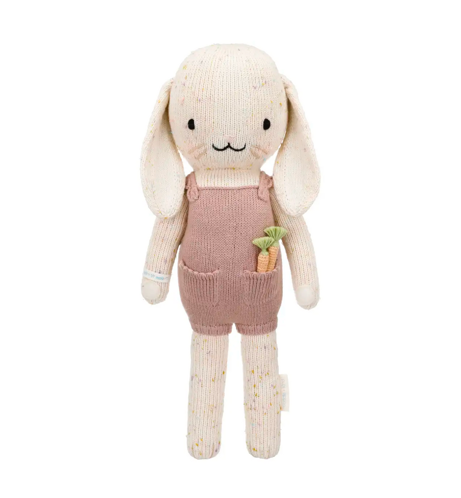 A Cuddle + Kind Bunny in a pink hand knit jumper and overalls, holding a small carrot, with a stitched face and long floppy ears, isolated on a white background.