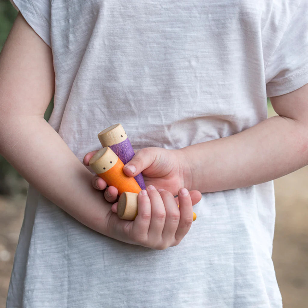 A child in a white t-shirt holds a small Grapat Sticks Gnomes toy figure in their hands, closely inspecting it. The background is softly blurred with natural tones.