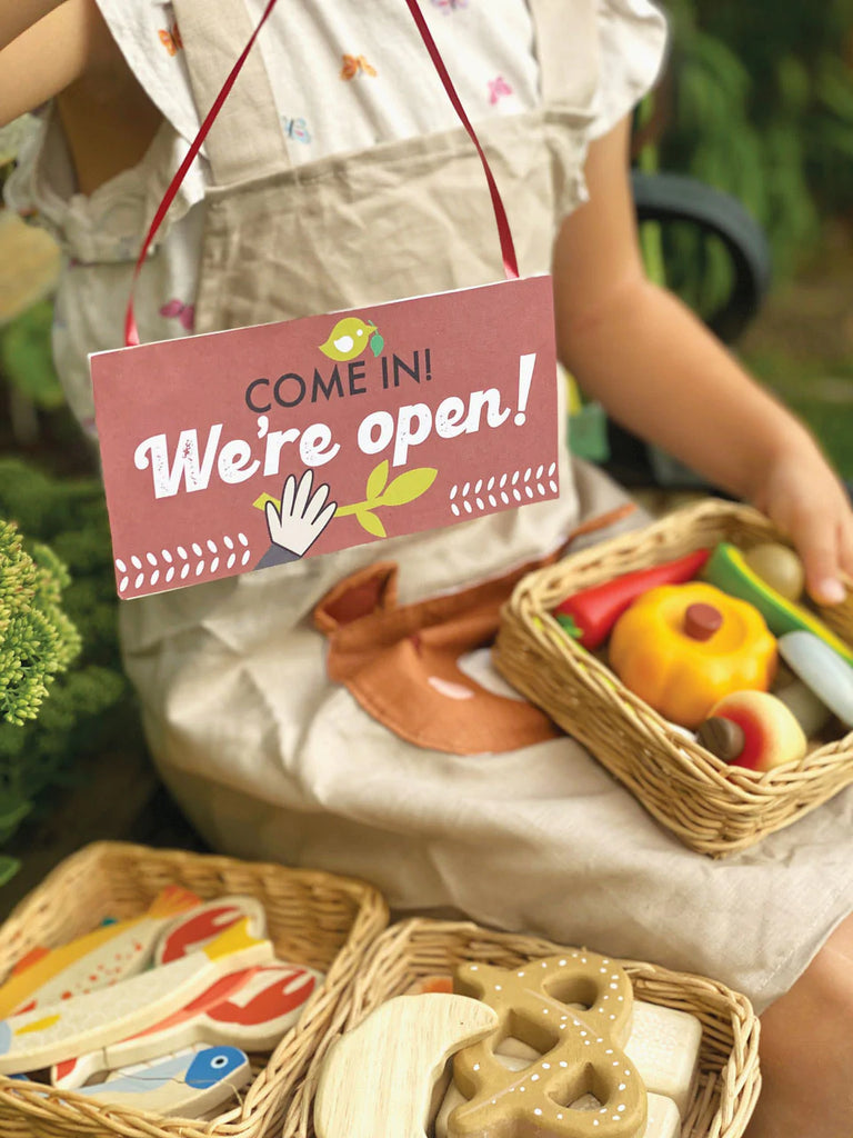A child holding a Market Basket full of colorful play fruits and vegetables in a garden with a "come in! we're open!" sign, suggesting an imaginative play of a market.