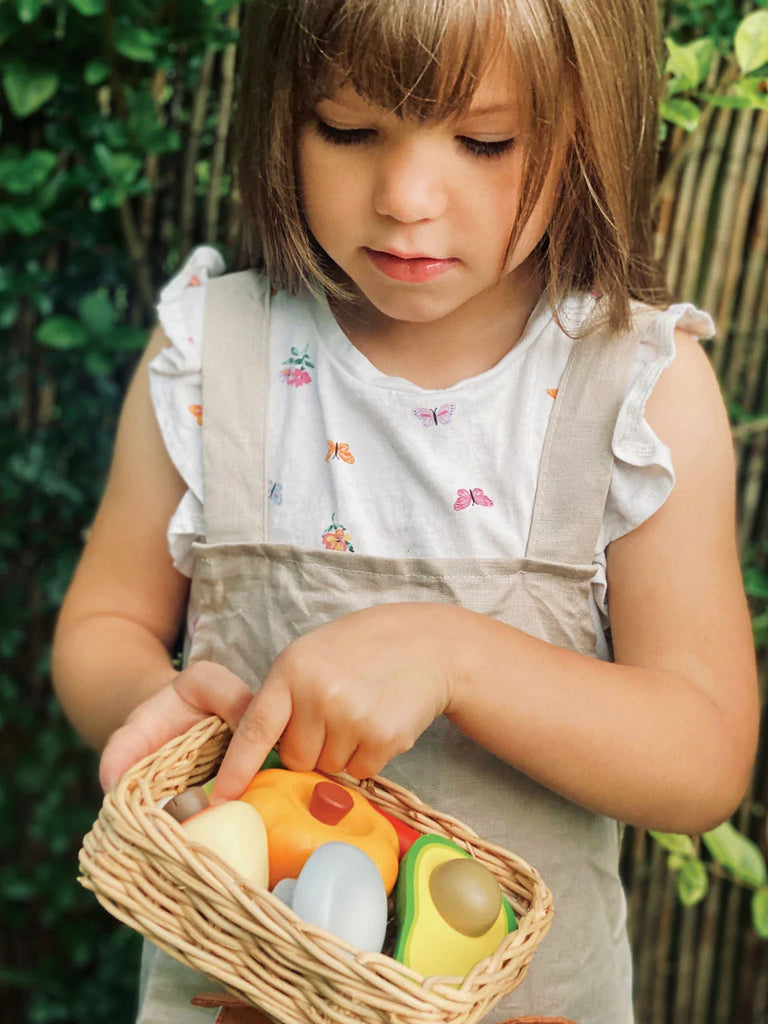 Sentence with product name: A young child holds a Market Basket filled with colorful eggs, intently examining them. She wears a light-colored dress with small embroidered details. The background features lush greenery.