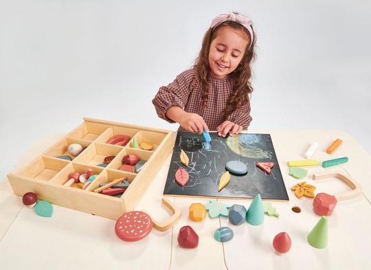 A young girl with a headband smiles while playing with a My Forest Floor full of colorful chalk and various geometrical shapes on a blackboard.