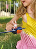 A young girl in a yellow and pink dress plays outdoors with a toy featuring Swifty Bird riding a blue and black airplane.