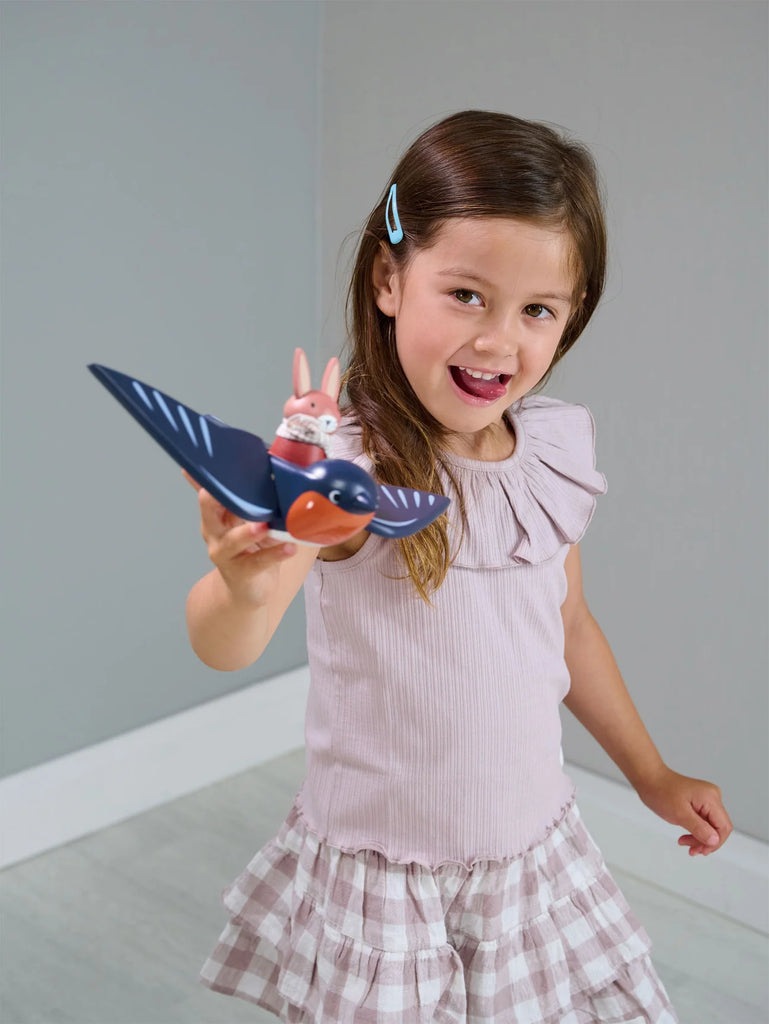 A young girl with brown hair, dressed in a pink ruffled top and plaid skirt, playfully holding a Swifty Bird towards the camera in a light gray room at Merrywood Village.