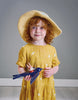 A young girl with curly red hair wearing a yellow dress and a straw hat, holding a blue toy airplane with "Swifty Bird" on it, standing in front of a gray background
