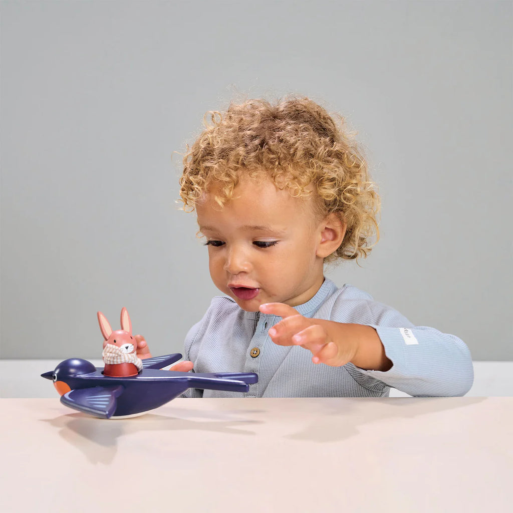 A toddler with curly hair is fascinated by a small Swifty Bird airplane and figures on a table, reaching out curiously with one hand. The background features illustrations of Merrywood Village.