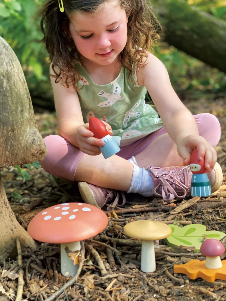 A young girl with curly hair sits cross-legged on wood chips, playing with the Woodland Gnome Family from the Merrywood Tales collection in a sun-dappled forest setting.