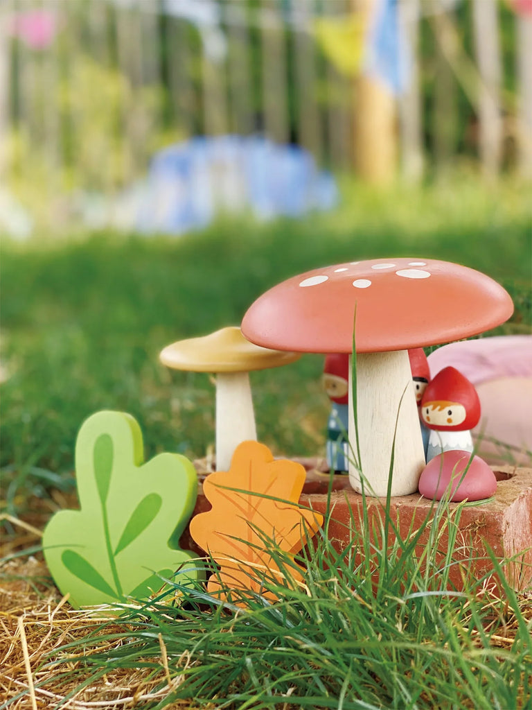 Decorative garden setting featuring ceramic mushrooms and colorful wooden leaves on pine straw, with a Woodland Gnome Family peeking from behind. Vibrant grass and blurred colorful fence in the background.