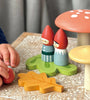 A child's hand is playing with a red toy piece beside wooden figurines of the Woodland Gnome Family characters and mushroom-shaped toys. The scene, reminiscent of the Merrywood Tales, is set on a patterned surface with additional toy elements like a yellow leaf.