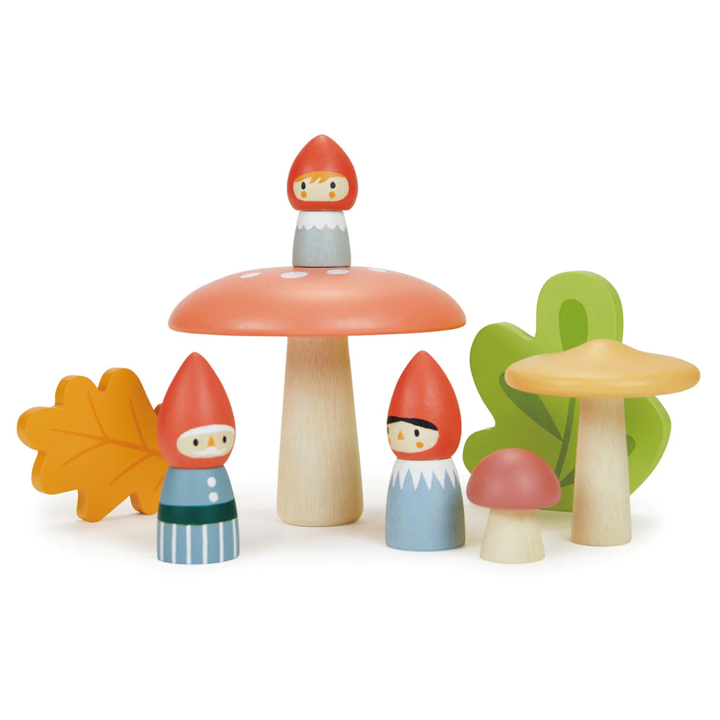 A whimsical scene from Merrywood Tales features the Woodland Gnome Family with red hats and painted clothes, surrounded by a variety of colorful wooden mushrooms, a yellow wooden leaf, and a green wooden plant. The gnome on top sits on the largest mushroom, overseeing the enchanted forest setting.