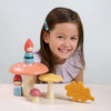 A young girl with a bright smile, looking at the camera, sits behind wooden toy mushrooms with small figures from the Woodland Gnome Family on them, alongside a wooden leaf.