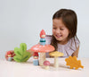 A young girl with long brown hair smiles while playing with colorful wooden toys from the Woodland Gnome Family collection. The toys include various mushroom shapes, figures in red hoods resembling Nordic gnomes, and leaf pieces, all arranged on a light-colored surface. The background is plain and light-colored.
