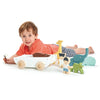 A toddler lying on their stomach next to The Friend Ship Pull Toy, holding a wooden figure, with a sustainable rubber wood animal set lined up nearby, on a white background.
