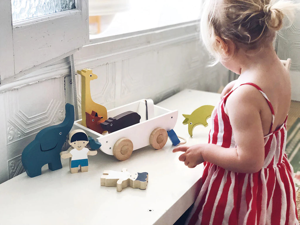 A young child in a striped dress plays with The Friend Ship Pull Toy and a character figurine next to a pull-along toy cart by a window.