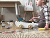 A toddler sitting on a patterned floor next to The Friend Ship Pull Toy filled with assorted toys, including a knitted bear and a pull-along toy elephant made from sustainable rubber wood, with cartoon cut