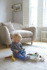 A young child sits on the floor playing with The Friend Ship Pull Toy next to a plush beige armchair in a brightly lit living room with white shutters.