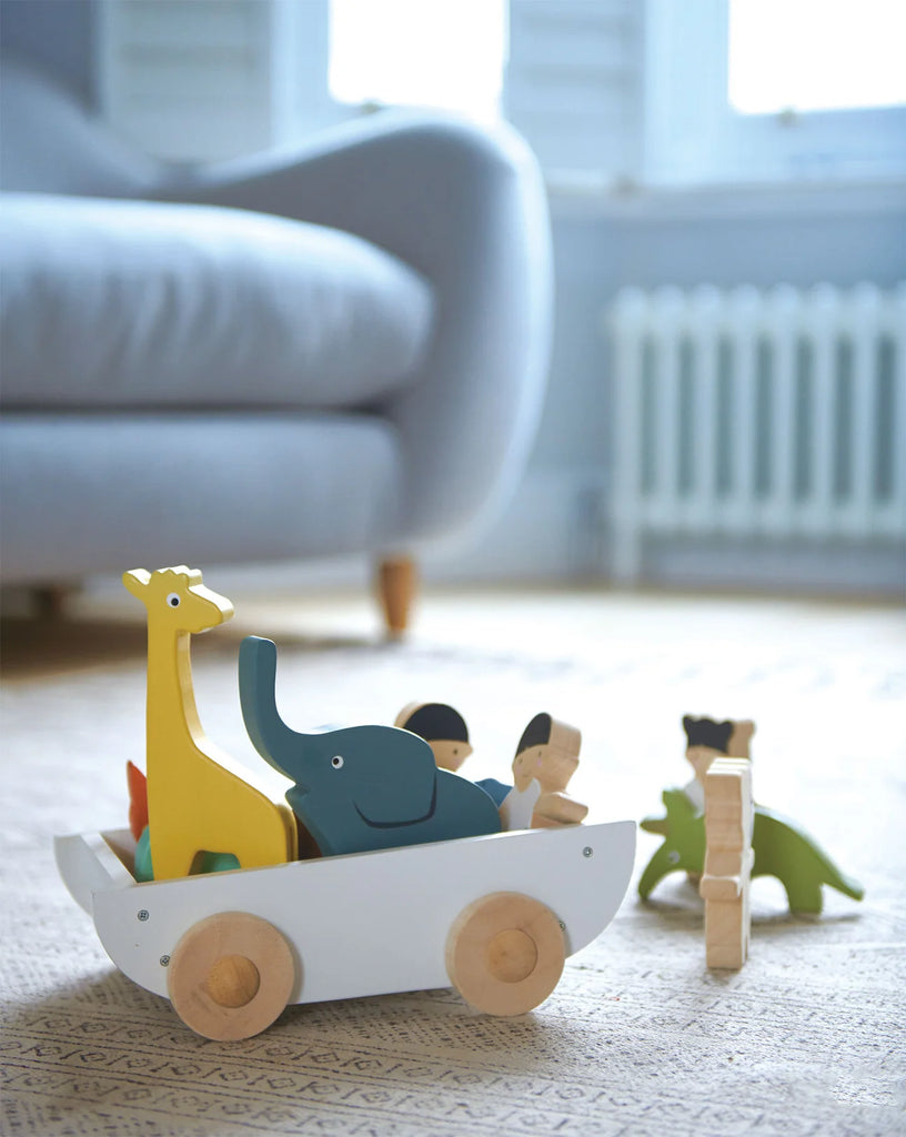 A colorful wooden Friend Ship Pull Toy with a giraffe, elephant, and other animals in a small wheeled cart on a carpeted floor, with a blurred background of a blue chair and a white heater.