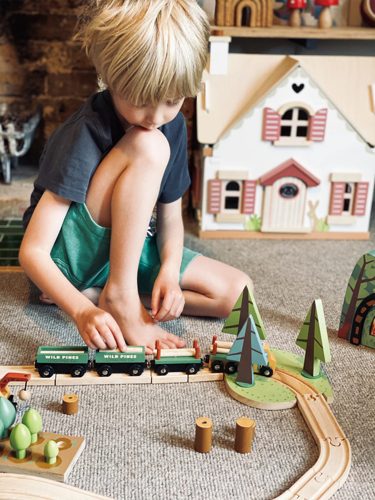 A young boy plays with a Wild Pines Train Set on a wooden floor, interacting with tracks and various props like trees and a house, focused and intrigued.