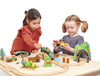 Two young girls playing with a Wild Pines Train Set that includes tracks, trees, buildings, and toy vehicles, interacting happily over their imaginative play setup.