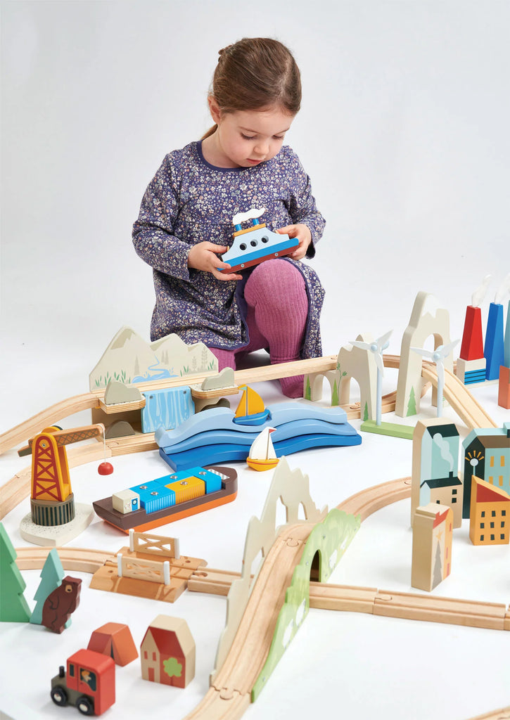 A young girl plays with a Mountain View Train set, arranging a blue train on elevated tracks surrounded by colorful wooden buildings, trees, and bridges on a white background.
