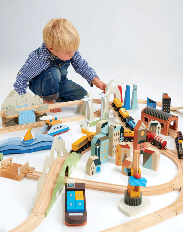 A young child plays with a Mountain View Train set, including wooden tracks, colorful buildings, and trains, on a white background.