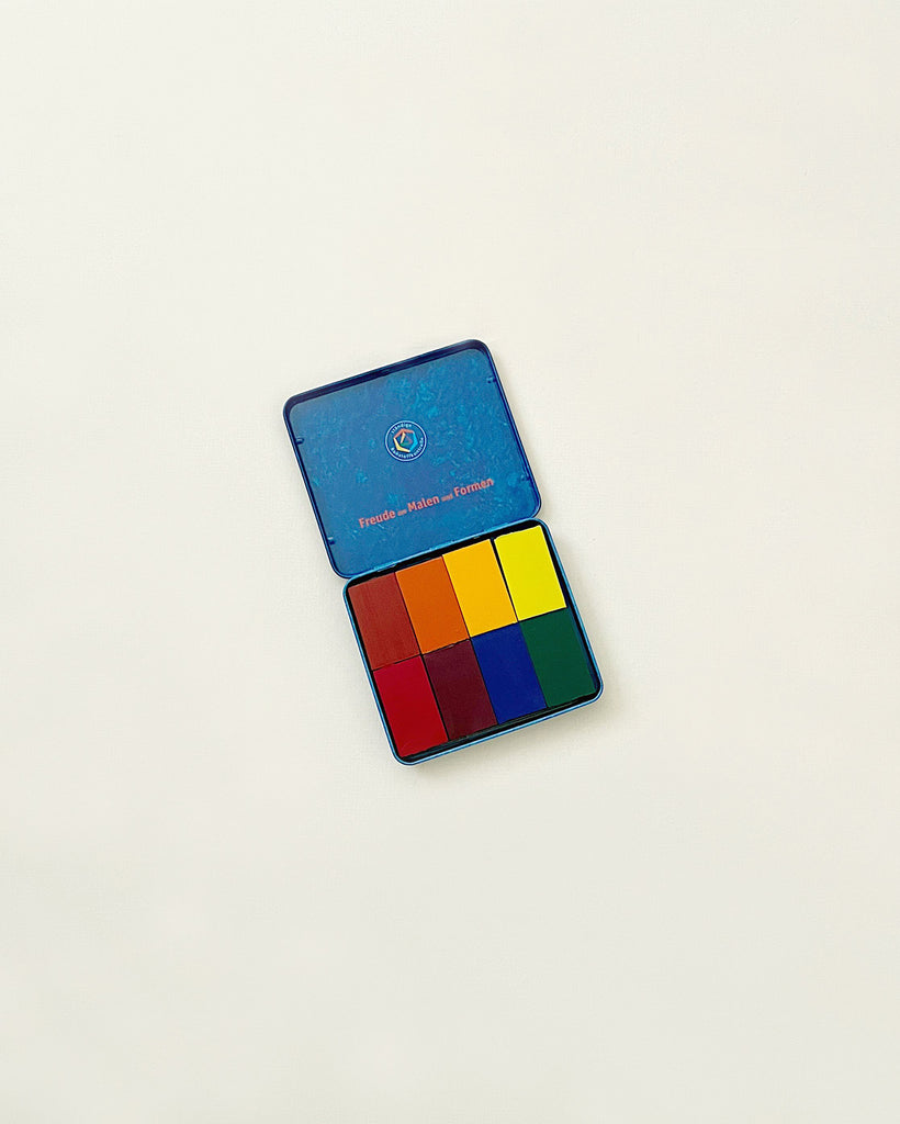 A blue Stockmar Wax Block Crayons Waldorf Tin Case with a colorful rectangle design resembling a pixelated image in Stockmar colors on the lower half, isolated on a white background.