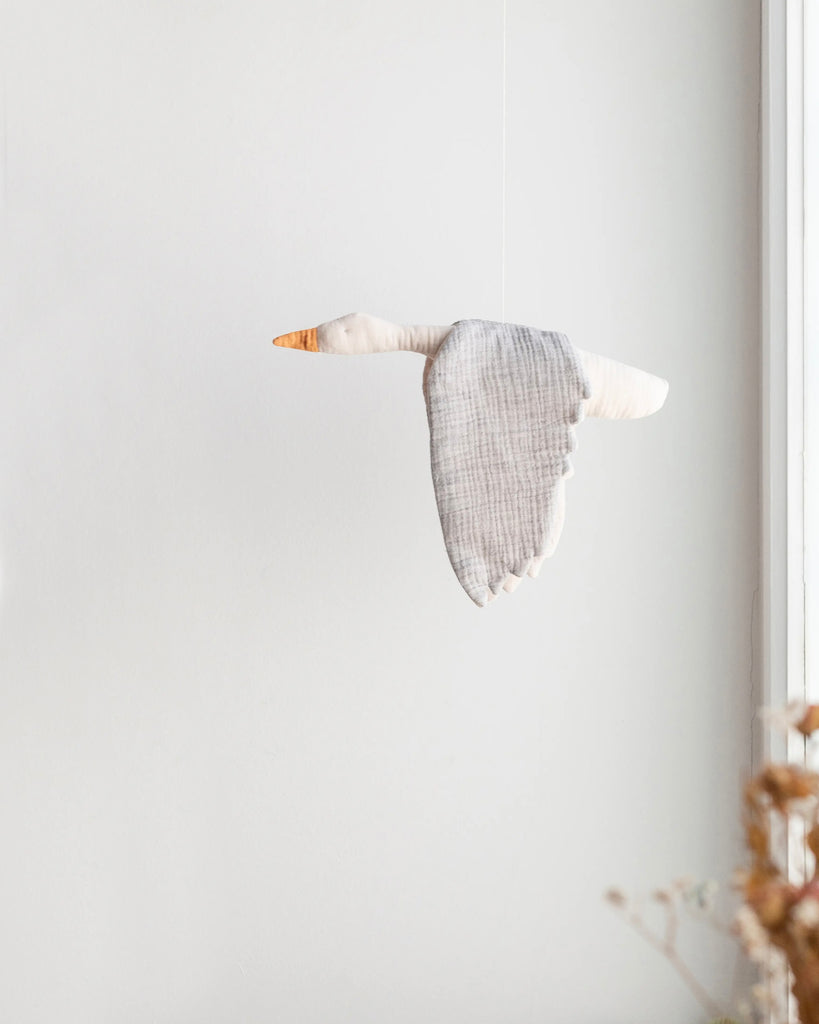 A Seagull Mobile, handmade in Spain, hangs against a light background near a window, casting a gentle shadow. The bird, made of plaid fabric, appears to be in mid-flight.