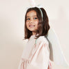 A young girl wearing a Meri Meri Winged Unicorn Costume set with pink iridescent sequin fabric smiles in a warmly lit studio setting.