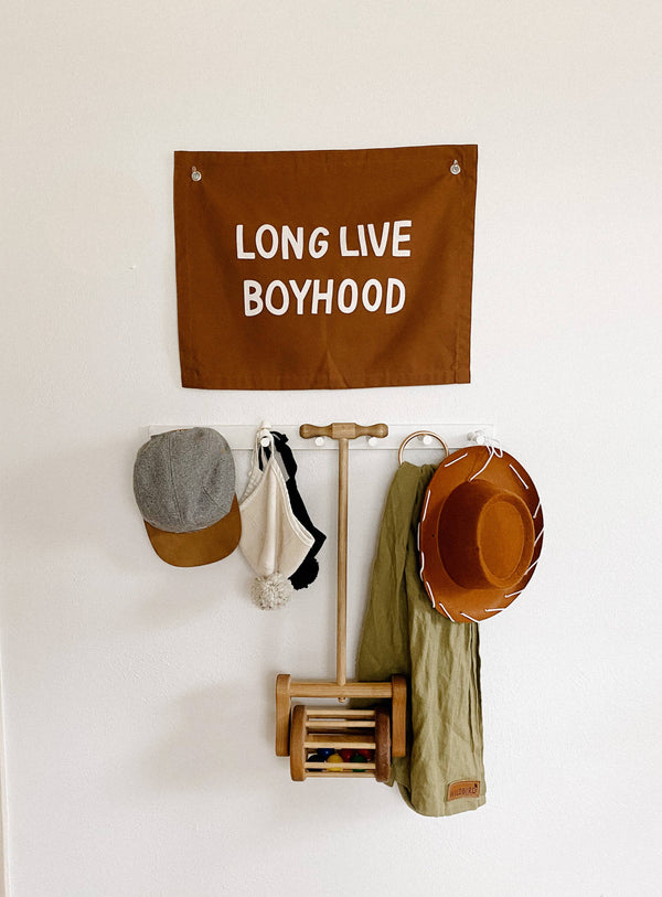 Long live boyhood banner in rust color on white wall. Items below banner are a gray and tan cap, white and black bonnets, wooden push toy, green army swaddle and tan hat. 