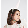 A young girl with shoulder-length brown hair, wearing a Meri Meri Winged Unicorn Costume with a pastel horn and ears, and a matching pale pink dress looks to her left.
