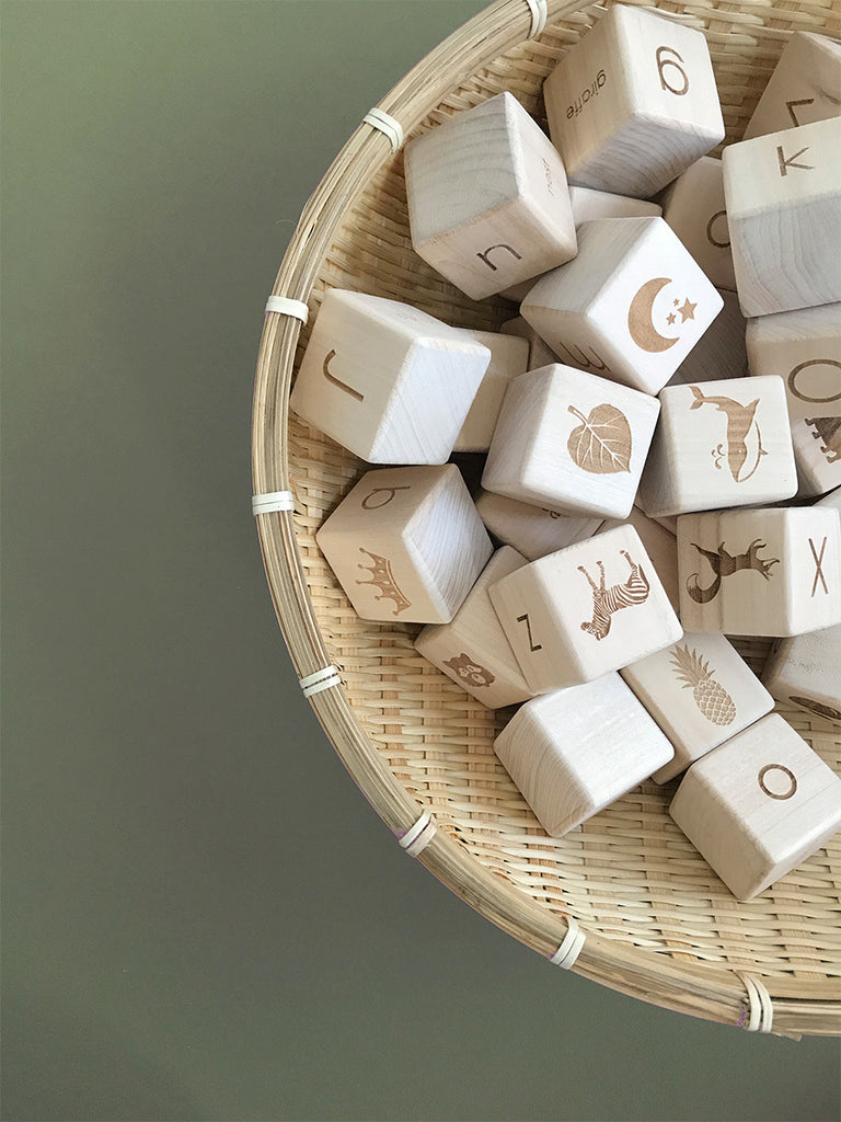 A collection of Alphabet Wooden Blocks made from natural linden wood with various letters and pictorial engravings, stored in a wicker basket on a grey background.