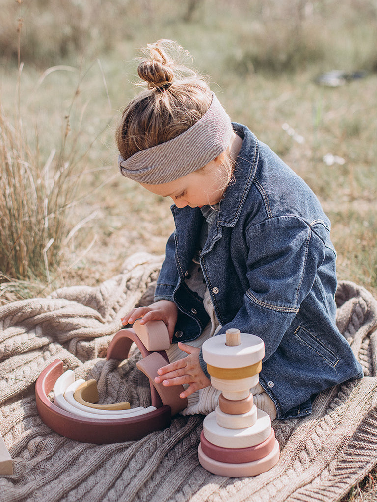 A young child wearing a denim jacket and headband plays with a Wooden Pyramid Stacker - Pink on a blanket outdoors, focusing intently on stacking colorful pieces.