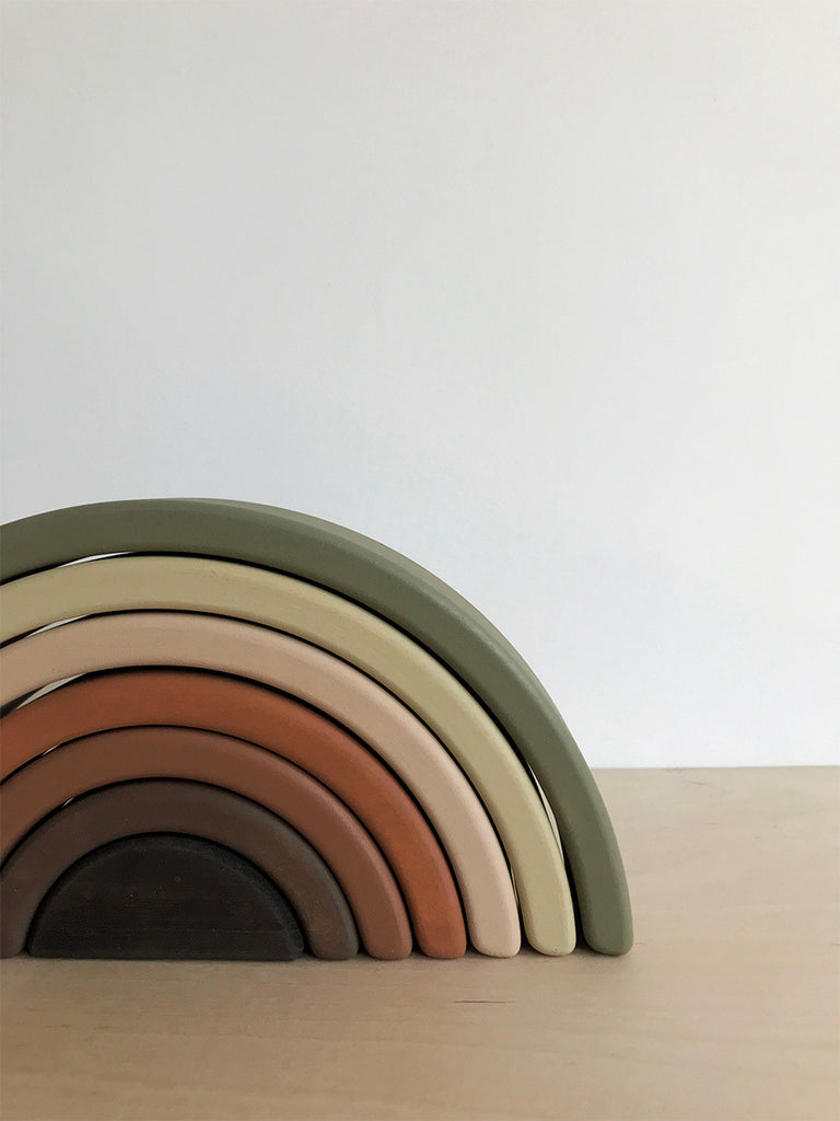 A Handmade Rainbow Stacker - Olive in various earthy tones (green, beige, tan, and brown). The arches, painted with non-toxic paint, are neatly nested within each other and positioned on a light wooden surface against a plain white background.