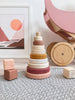 A stylish interior scene featuring a Wooden Pyramid Stacker - Pink on a carpet, designed to enhance hand-eye coordination, with soft pink and neutral tones. Artistic wall art and a handmade wooden toy cart are included.
