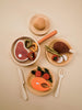 Flat lay of Sabo Concept Handmade Wooden Dinner Set arranged on circular plates: meats, vegetables, fruits, and eggs, with utensils and bowls, all on a soft beige background.