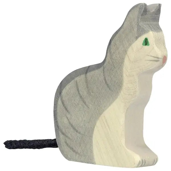 A small, wooden Holztiger Sitting Gray Cat with gray stripes and white fur. The cat is sitting upright with its tail lying flat on the ground. The carved features give it a simple, handcrafted appearance, reminiscent of high-quality HOLZTIGER figures, with green eyes and a pink nose.