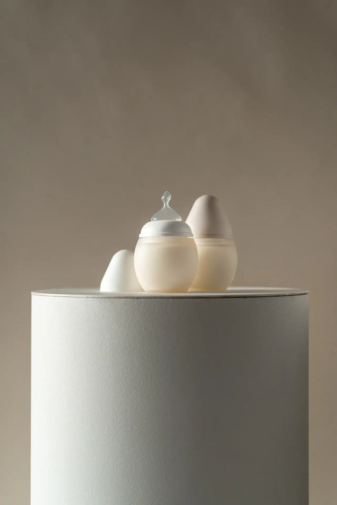 Two eggs with a drop-shaped medical grade silicone baby bottle cap on top, one larger and one smaller, are displayed on a cylindrical gray podium against a soft beige background.