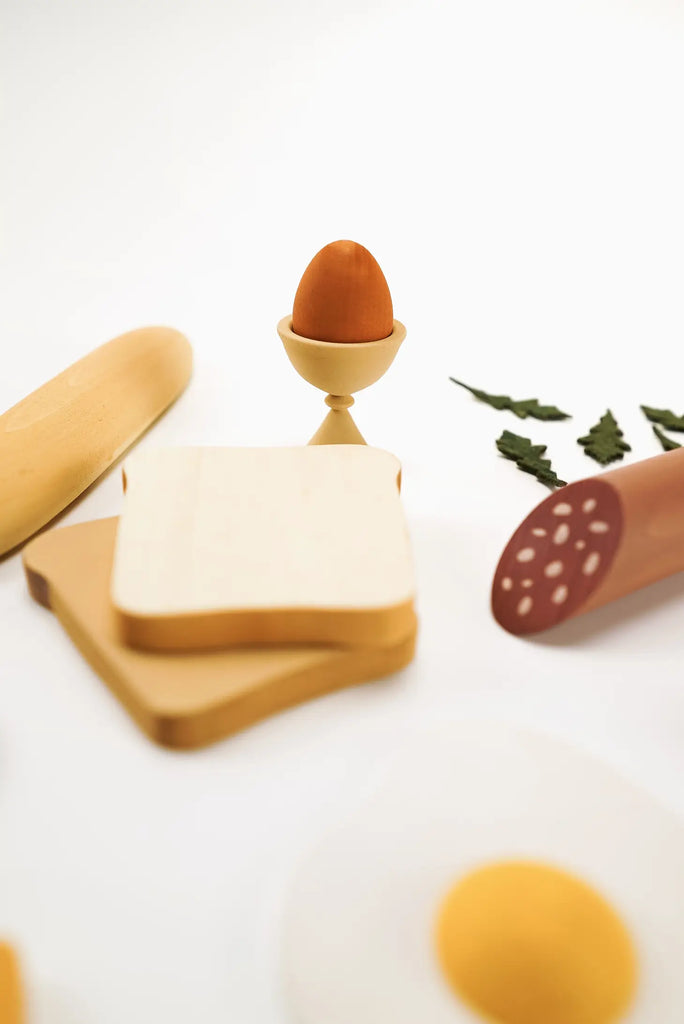 A Sabo Concept Handmade Wooden Breakfast Set, consisting of a boiled egg in a wooden egg cup, with a stack of cheese slices, rolling pin, and a wooden spoon with leaf decorations nearby, all crafted from non-toxic water-based paint.