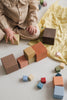 A toddler in a checkered outfit sits on the floor, reaching out towards the Raduga Grez Big Cube Block Set scattered around on a pale yellow crinkled sheet.
