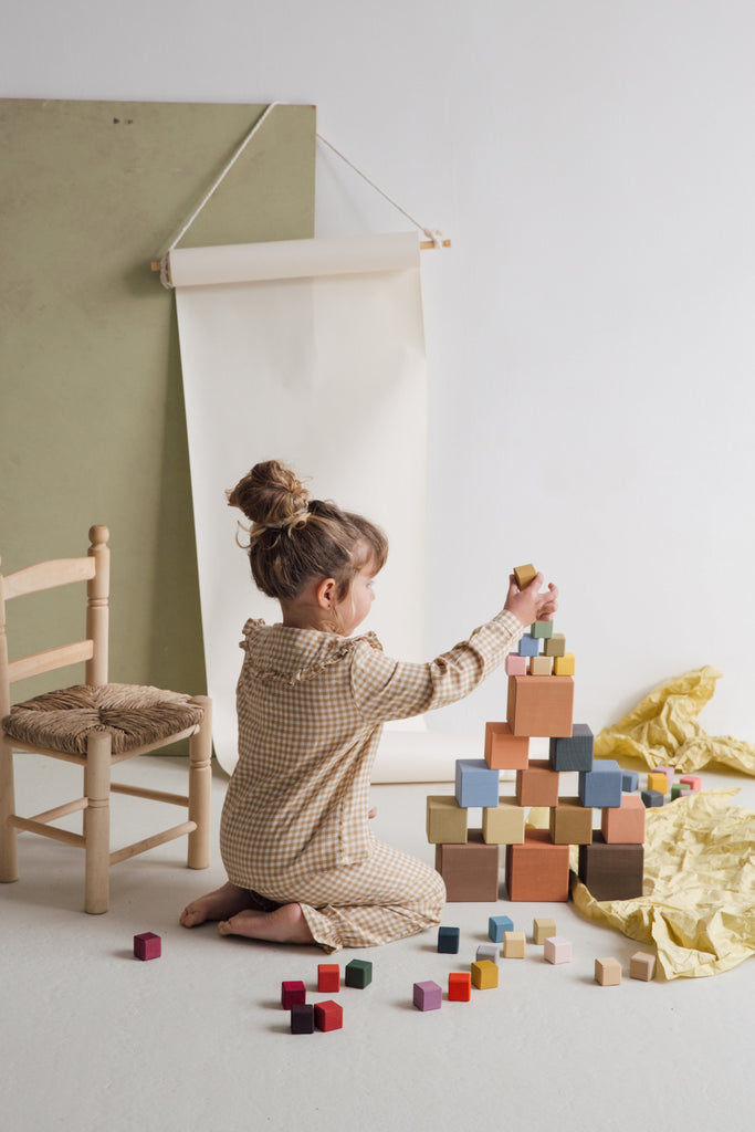 A young child with a bun hairstyle is seated on the floor, building a tall tower with Raduga Grez Big Cube Block Set in a studio-like setting with a white backdrop and a wooden chair.