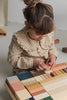 A young child wearing a brown and white checkered outfit is playing with a Raduga Grez Extra Large Building Blocks Set, focusing intently on arranging the assortment of colorful, hand-crafted wooden blocks coated in non-toxic water-based paint. The setting appears to be a clean, minimalist room with a light-colored floor.
