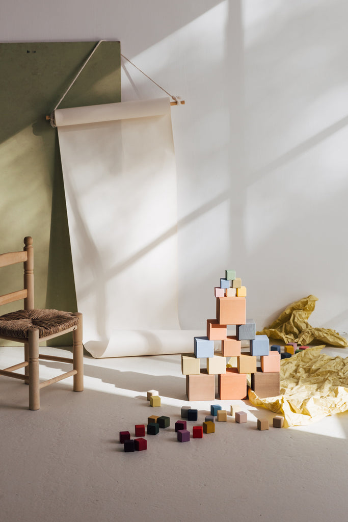 A minimalist room with soft sunlight casting shadows on the wall and floor, featuring a wooden chair and a Raduga Grez | Big Cube Block Set with scattered pieces around.